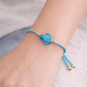 May include: A turquoise adjustable bracelet featuring a small, detailed blue fish charm with a red eye. The bracelet has a woven cord and gold-coloured accents. The fish charm is approximately 2 cm long.