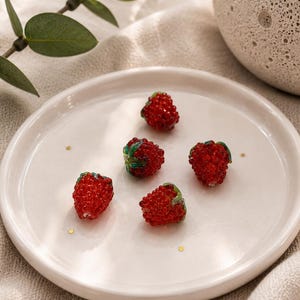Könnte beinhalten: Fünf kleine, rote, texturierte Glasperlen in Himbeerform, jede mit winzigen grünen Glasblättern an der Oberseite. Die Perlen sind auf einem glatten, runden, cremeweißen Keramikteller mit erhöhtem Rand angeordnet, der mit kleinen goldenen Sprenkeln versehen ist.
