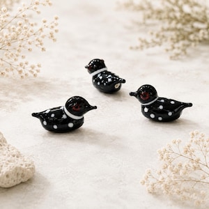 Puede incluir: Tres figuras decorativas de vidrio de somorgujo. Cada somorgujo es negro con lunares blancos y un collar blanco. Los somorgujos tienen ojos rojos y est&aacute;n dispuestos sobre una superficie neutra con flores secas.