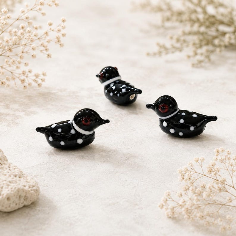 Puede incluir: Tres figuras decorativas de vidrio de somorgujo. Cada somorgujo es negro con lunares blancos y un collar blanco. Los somorgujos tienen ojos rojos y est&aacute;n dispuestos sobre una superficie neutra con flores secas.