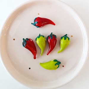 May include: A selection of glass chili peppers in red and green, with green stems, presented on a pale pink plate. The peppers have a glossy finish and appear handmade. The plate has a simple, minimalist design.
