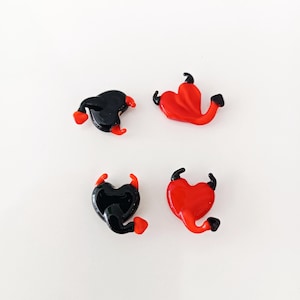May include: Four heart-shaped glass ornaments. Two are black with red accents, and two are red with black accents. Each ornament has small horns and a tail, suggesting a devilish theme. The ornaments are displayed on a white surface.