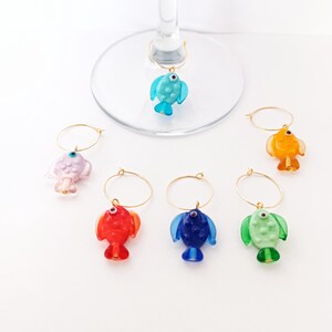 May include: A collection of colorful fish-shaped wine charms. Each charm is a different color, including blue, red, orange, green, and pink, with gold-colored hoops. The charms are designed to hang on the stem of a wine glass.