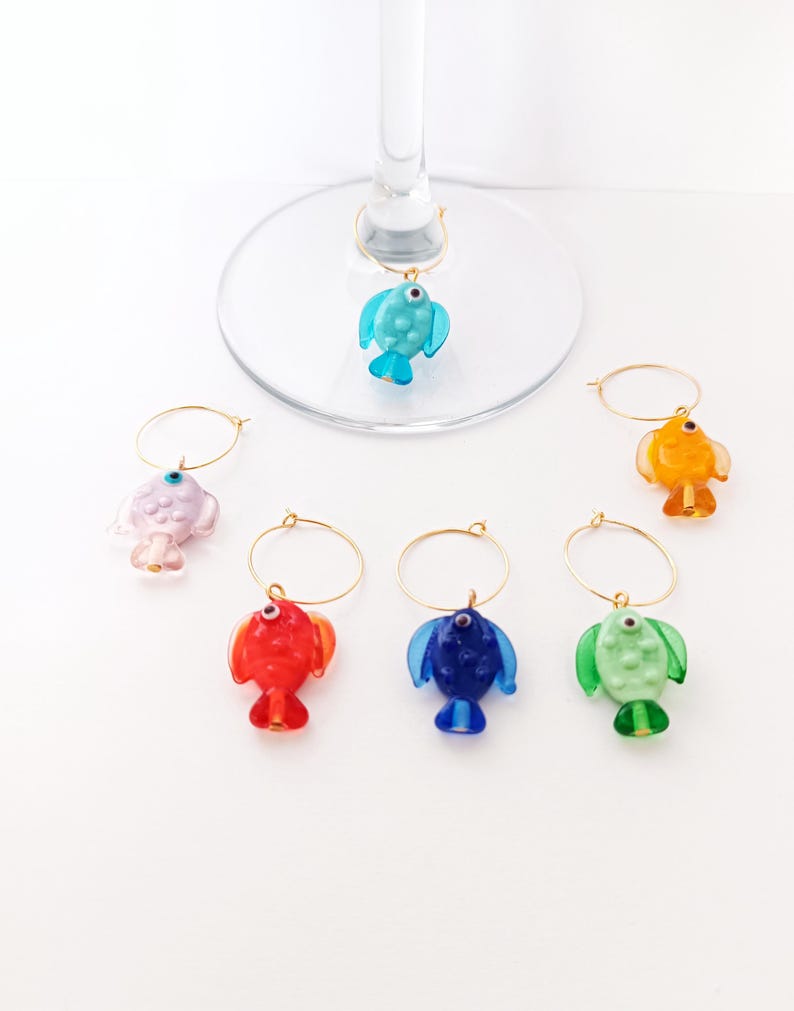 May include: A collection of colorful fish-shaped wine charms. Each charm is a different color, including blue, red, orange, green, and pink, with gold-colored hoops. The charms are designed to hang on the stem of a wine glass.