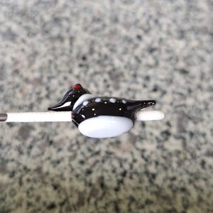 Puede incluir: Una figura de pato de vidrio hecha a mano. El pato presenta un cuerpo negro con lunares blancos, una barriga blanca y una cabeza roja. Est&aacute; montado sobre un palito blanco, mostrando los intrincados detalles del trabajo en vidrio.