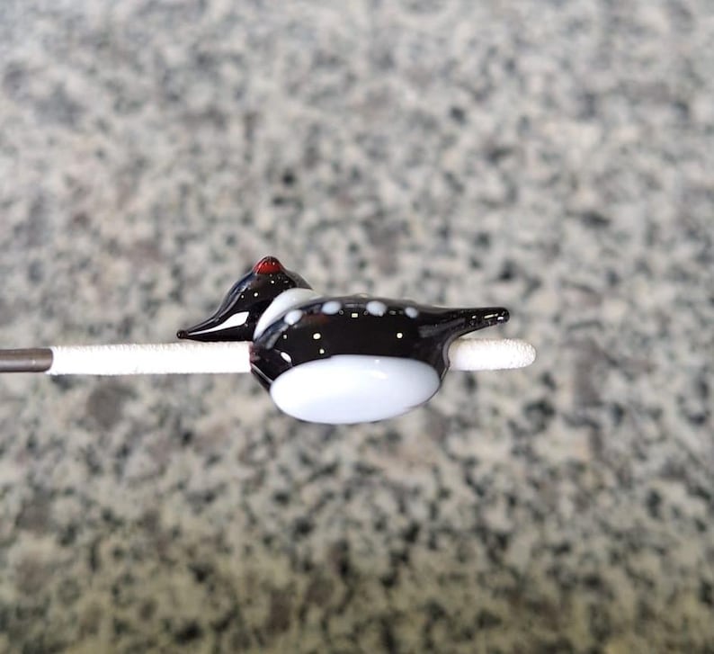Puede incluir: Una figura de pato de vidrio hecha a mano. El pato presenta un cuerpo negro con lunares blancos, una barriga blanca y una cabeza roja. Est&aacute; montado sobre un palito blanco, mostrando los intrincados detalles del trabajo en vidrio.