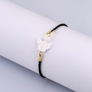 以下が含まれることがあります： 黒い紐のブレスレットに、白いセラミック製の天使のチャームが付いています。チャームには金色のアクセントがあります。