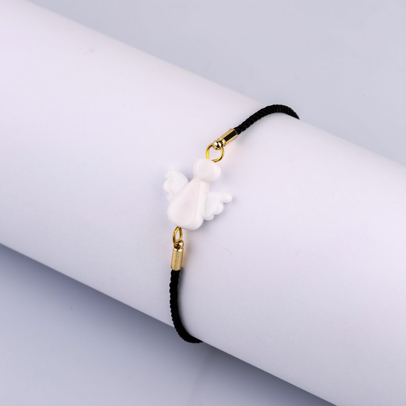 以下が含まれることがあります： 黒い紐のブレスレットに、白いセラミック製の天使のチャームが付いています。チャームには金色のアクセントがあります。