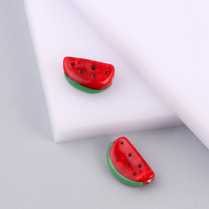 May include: Two small, crescent-shaped beads designed to resemble watermelon slices. Each bead features a glossy red top section with small black dots representing seeds and a smooth green bottom edge, crafted with a polished, reflective finish.