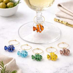 May include: A wine glass with a fish-shaped charm. Six additional fish charms in blue, light blue, green, yellow, and brown are displayed. The fish charms are attached to gold-coloured rings. Olives and rosemary sprigs are in the background.