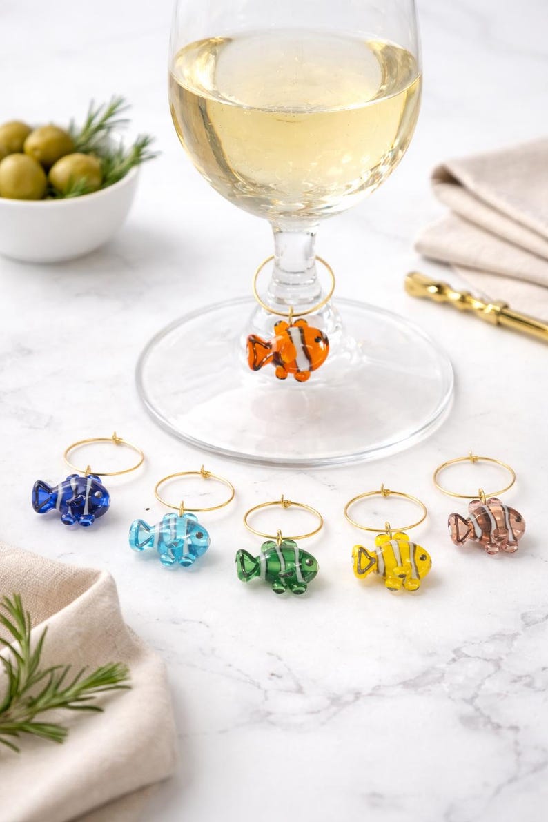 K&ouml;nnte beinhalten: Ein Weinglas mit einem bunten fischf&ouml;rmigen Charm. Sechs weitere Fisch-Charms in Blau, Hellblau, Gr&uuml;n, Gelb und Braun werden gezeigt. Die Fisch-Charms sind an goldfarbenen Ringen befestigt. Oliven und Rosmarinzweige im Hintergrund.