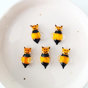 May include: Five small, glass bee figurines with yellow and black bodies, clear wings, and smiling faces. The bees are arranged on a white, round plate. The figurines are approximately 1 inch in length.
