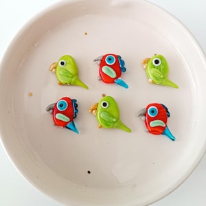 Op de afbeelding: Zes kleurrijke glazen papegaai-beeldjes. De papegaaien zijn in tinten groen en rood, met blauwe en gele accenten. Ze zijn gerangschikt op een witte, ronde plaat. Elke papegaai heeft een snavel, oog en staart.