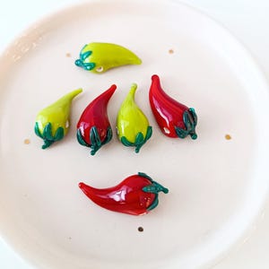 May include: Glass chili pepper ornaments in red and green, displayed on a white plate. The peppers have green stems. These are likely decorative items, perhaps for a kitchen or dining setting.