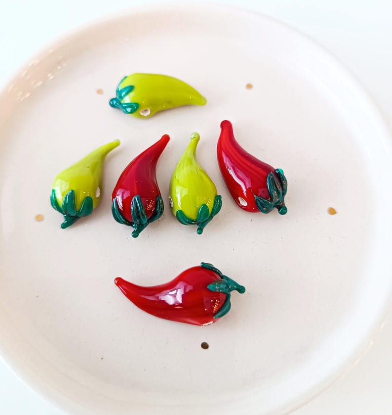 May include: Glass chili pepper ornaments in red and green, displayed on a white plate. The peppers have green stems. These are likely decorative items, perhaps for a kitchen or dining setting.