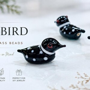 Puede incluir: Tres cuentas de vidrio negro y blanco con forma de somorgujo, con lunares blancos y ojos rojos. El texto "LOON BIRD" y "LAMPWORK GLASS BEADS" se muestra encima de los p&aacute;jaros. Tambi&eacute;n es visible la frase "Handmade with Nature in Mind".