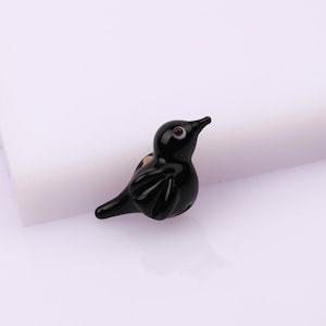 May include: A small black glass bird figurine with a white eye and a brown iris. The bird is perched on a white surface.