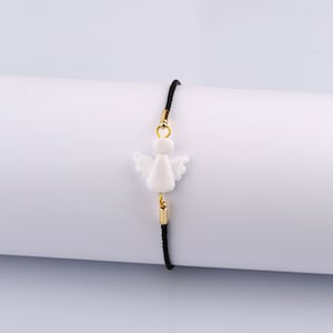 以下が含まれることがあります： 黒い紐のブレスレットに、白い天使のチャームと金のアクセントが付いている。