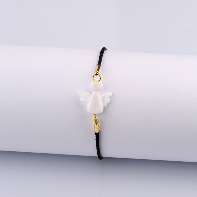 以下が含まれることがあります： 黒い紐のブレスレットに、白い天使のチャームと金のアクセントが付いている。