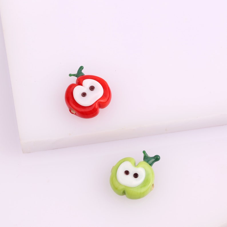 May include: Two glass apple beads, one red and one green, with white centers and black eyes. The beads are perfect for jewelry making or crafting.