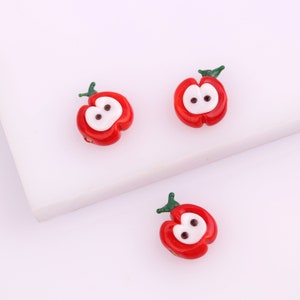 May include: Three red glass apple beads with white centers and black eyes. The beads have green stems.