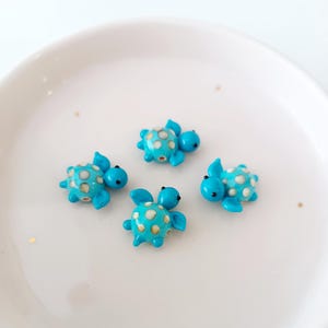 May include: Four small, turquoise turtle figurines with white polka dots and black eyes are arranged on a white surface. The turtles have flipper-like appendages and are facing different directions.