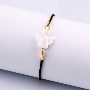 以下が含まれることがあります： 黒い紐のブレスレットに、白いセラミック製の天使のチャームが付いています。チャームには金色のアクセントがあります。