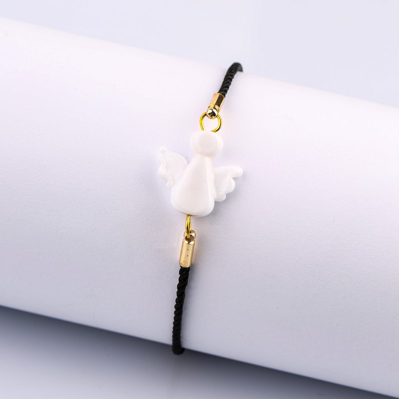 以下が含まれることがあります： 黒い紐のブレスレットに、白いセラミック製の天使のチャームが付いています。チャームには金色のアクセントがあります。