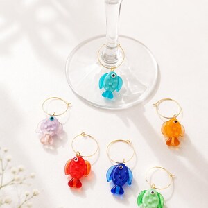 May include: Set of six colorful fish-shaped wine charms. Each charm features a different color: blue, purple, red, orange, blue, and green. The fish are attached to gold-colored metal rings, designed to hang on the stem of a wine glass.