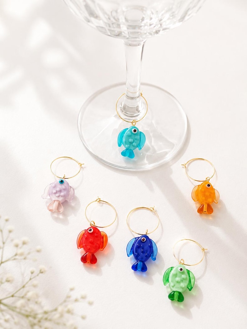 May include: Set of six colorful fish-shaped wine charms. Each charm features a different color: blue, purple, red, orange, blue, and green. The fish are attached to gold-colored metal rings, designed to hang on the stem of a wine glass.