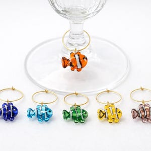 K&ouml;nnte beinhalten: Eine Sammlung farbenfroher Weincharm-Anh&auml;nger in Fischform. Jeder Anh&auml;nger besteht aus Glas und weist ein anderes Farbschema auf, darunter Orange, Blau, Gr&uuml;n, Gelb und Rosa. Die Anh&auml;nger h&auml;ngen an goldfarbenen Metallringen.