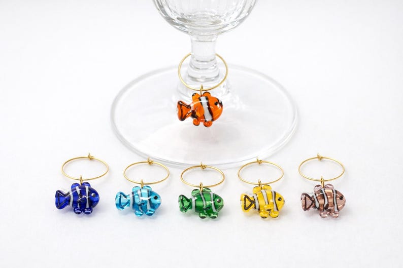 K&ouml;nnte beinhalten: Eine Sammlung farbenfroher Weincharm-Anh&auml;nger in Fischform. Jeder Anh&auml;nger besteht aus Glas und weist ein anderes Farbschema auf, darunter Orange, Blau, Gr&uuml;n, Gelb und Rosa. Die Anh&auml;nger h&auml;ngen an goldfarbenen Metallringen.