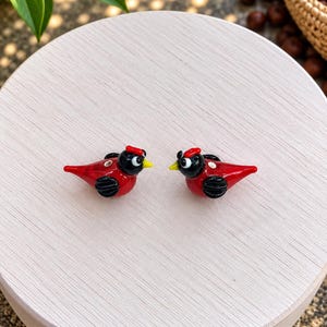 Op de afbeelding: Twee kleine glazen vogelbeeldjes met felrode lichamen, zwarte koppen en zwarte vleugels. Elke vogel heeft een kleine gele snavel, een wit oog met een zwarte pupil en een piepkleine rode kuif boven op de kop.
