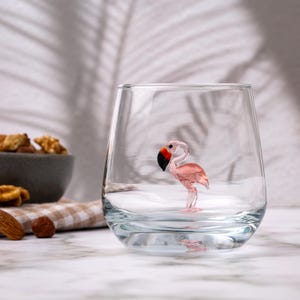May include: A clear glass tumbler with a pink flamingo figurine inside. The flamingo is made of glass and has a red beak and black eyes.