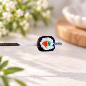 Op de afbeelding: Een haarstokje met een decoratieve glazen kraal in de vorm van een sushirol. De kraal heeft een zwarte buitenlaag, een witte rijstkern en een klein oranje en groen accent in het midden. Het stokje zelf is dun, recht en donker van kleur.