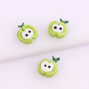 May include: Three green glass apple beads with white centers and brown eyes. The beads have a green stem and leaf on top.