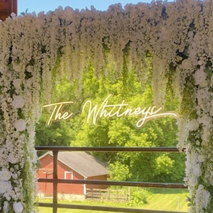May include: A white floral archway with a neon sign that reads "The Whitneys" in a rustic barn setting.