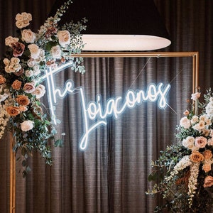 May include: A gold frame with a white neon sign that reads "The Joiaconos" is decorated with two large floral arrangements. The flowers are a mix of white, pink, and peach roses, and greenery. The frame is set against a backdrop of dark gray curtains.