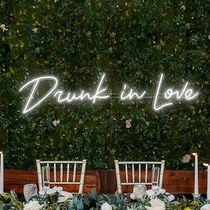 Puede incluir: Una mesa de boda con velas blancas, vegetación y flores blancas. El fondo es una pared de musgo verde con un letrero de neón blanco que dice "Drunk in Love."
