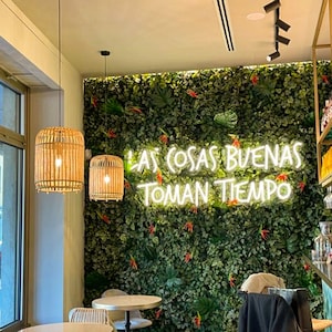 Può includere: Interno di un bar con un muro verde e l'insegna al neon "LAS COSAS BUENAS TOMAN TIEMPO". Due lampade a sospensione in tessuto sono appese sopra piccoli tavoli rotondi e sedie. Il bar ha un'estetica moderna e naturale.