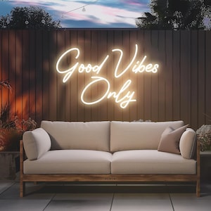 May include: White neon sign with the text "Good Vibes Only" mounted on a brown wooden wall. A beige couch with a light brown wooden frame sits in front of the sign.
