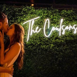 May include: A couple kisses in front of a green wall with a white neon sign that reads "The Coufsons".