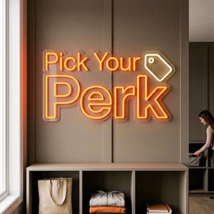 Könnte beinhalten: Ein orangefarbenes Neon-Schild, 76 cm breit, mit den Worten "Pick Your Perk" und einem weißen Etikett-Symbol. Darunter befindet sich ein Regal mit einer Tragetasche, gefalteter Kleidung und Notizbüchern. Das Schild ist an einer Wand befestigt.