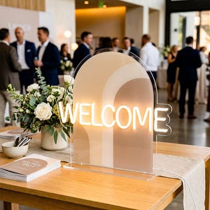 Pode incluir: Uma placa de néon branco quente "WELCOME", com cerca de 51 cm de altura, com um fundo em camadas bege e creme. A placa é exibida numa mesa de madeira com um corredor de tons neutros, com um arranjo floral e uma pilha de livros.