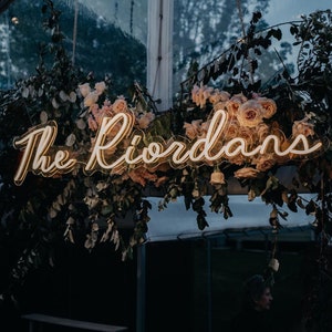 K&ouml;nnte beinhalten: Ein Neon-Schild mit dem Text "The Riordans" in wei&szlig;er Schrift, das an einem Blumenarrangement aus rosa Rosen und Gr&uuml;n h&auml;ngt.