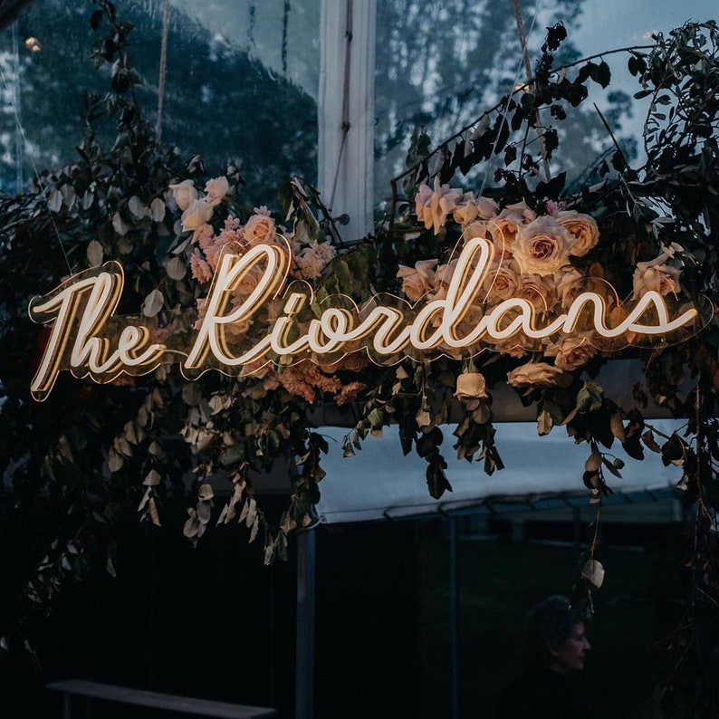 K&ouml;nnte beinhalten: Ein Neon-Schild mit dem Text "The Riordans" in wei&szlig;er Schrift, das an einem Blumenarrangement aus rosa Rosen und Gr&uuml;n h&auml;ngt.