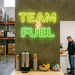 May include: A neon sign reading "TEAM FUEL" in lime green and white, with a lightning bolt symbol. A coffee maker, paper cups, and a bowl of fruit are on a wooden counter. The sign is 91 cm wide.