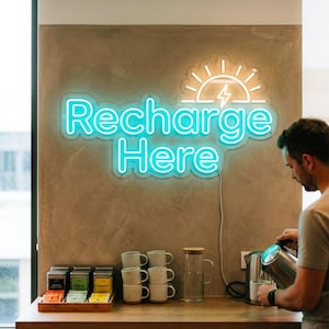 May include: A neon sign with the words "Recharge Here" in ice blue, with a sun and lightning bolt design. The sign is 36 inches wide and has warm white accents. The sign is mounted on a textured wall above a wooden counter with tea and mugs.