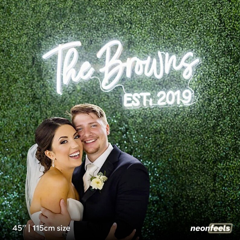 May include: A bride and groom pose in front of a green wall with a white neon sign that reads "The Browns Est. 2019" and measures 45 inches or 115 centimeters.