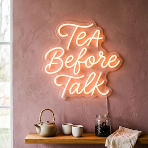 May include: A warm white neon sign that reads "Tea Before Talk" is mounted on a textured pink wall. Below, a wooden shelf holds a teapot, two white cups, a jar, and a folded cloth. The sign is 20 inches in size.